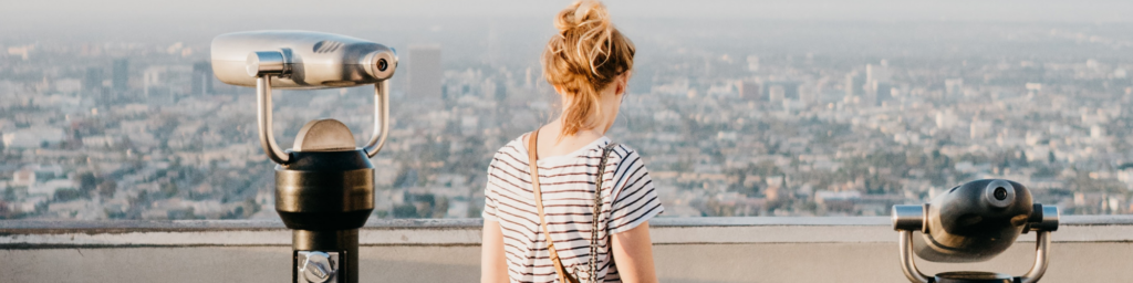 Woman pictured, standing to the right of an eye viewing machine overlooking a beautiful landscape, wearing a black and white striped top