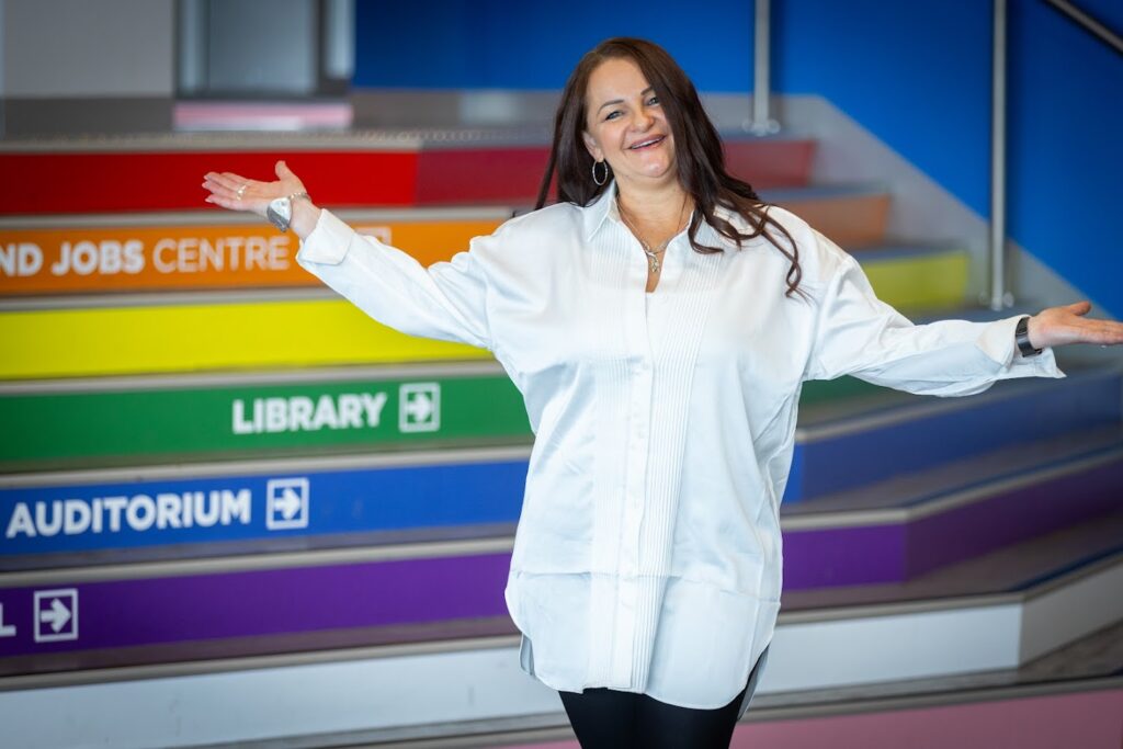 Pictured, Heather Thompson with a big smile on her face with her hands up in the air. Wearing a white shirt and beautiful silver jewelry, her long dark hair comes over the front of her white shirt. In the background are bright coloured stairs in purple, blue, green, yellow, orange and red.