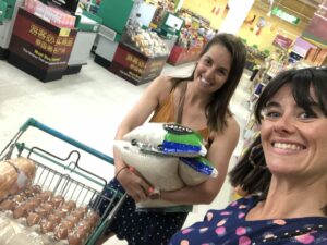 Donna Lindsay and Phoebe Wilkens in Thailand wearing summer clothes for the hot weather standing in front of the camera taking a selfie with supplies in the background, Donna is holding three big bags of rice and Phoebe is pushing a trolley full of eggs and other supplies