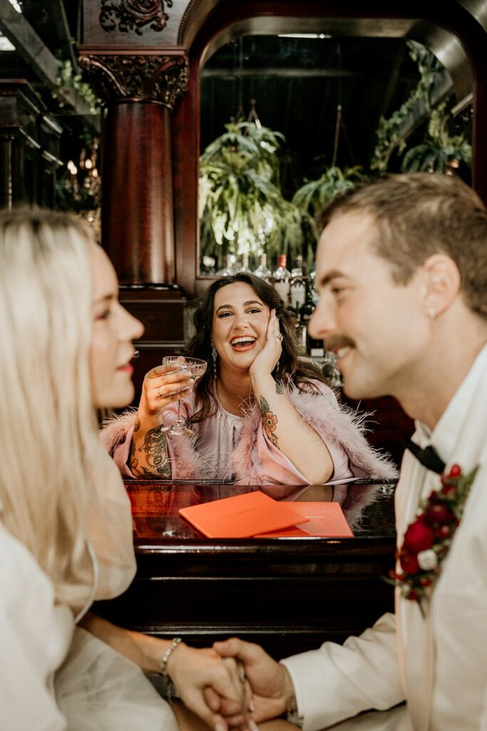 Pictured: Maddison Gillespie, sitting smiling with a cocktail glass in one hand and her other hand on her face. In front of her is a newly married couple.