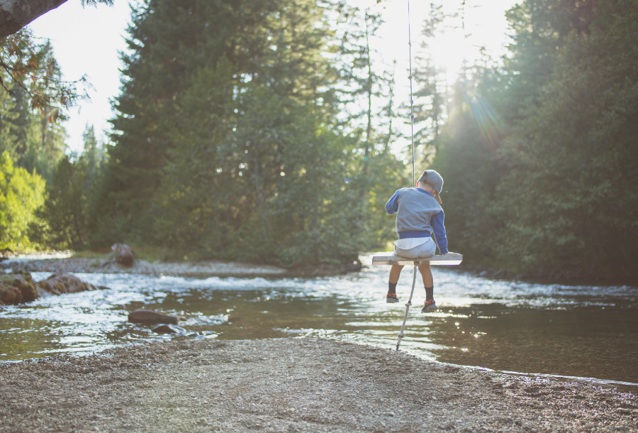 Picture of a child on a tree swing with his head bowed over the bank of a running river. Large pine trees with the sun shining through them are in the background of the picture