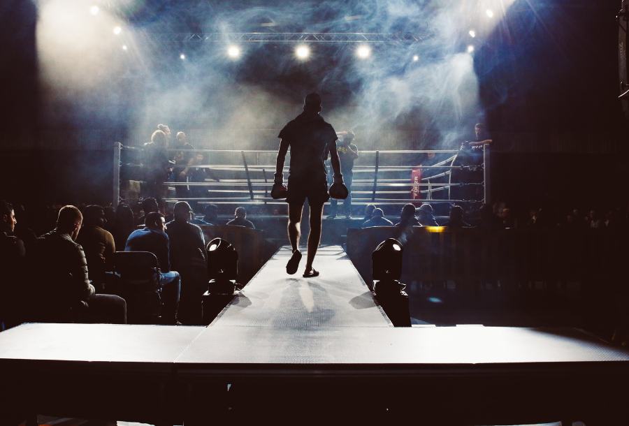 Picture of a man walking away from the centre of the screen down towards a boxing ring, wearing boxing gloves and dark clothes, his head is bowed and the spotlights are on him as he is drawing closer to the boxing ring