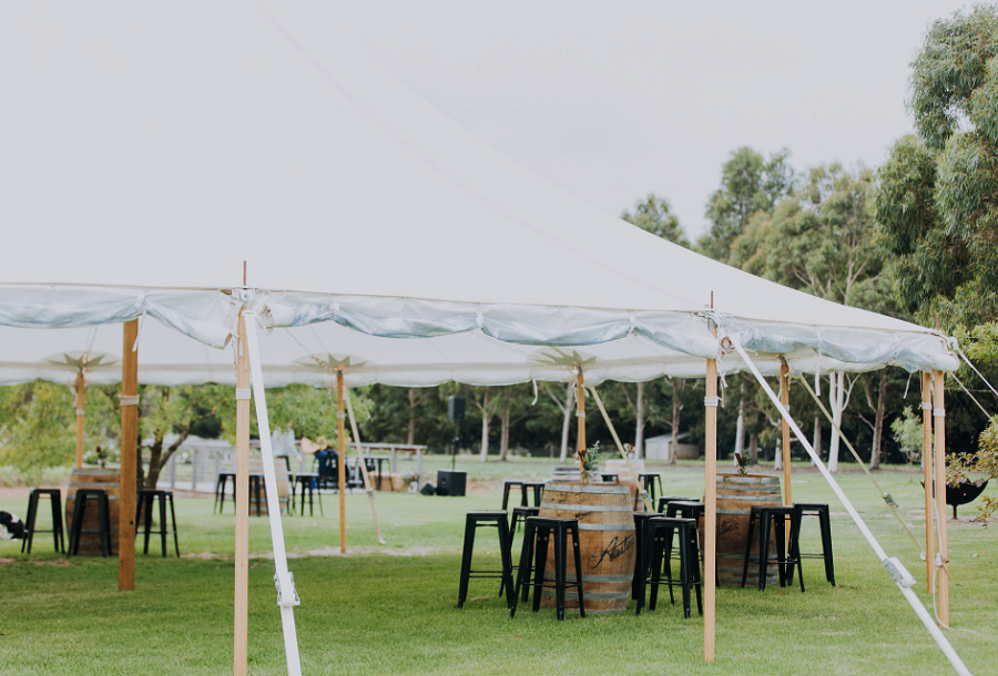 IMAGE DESCRIPTION: Big white tee-pee shaped tent on green grass in an open backward, surrounded by wine barrels being used for tables and black stools