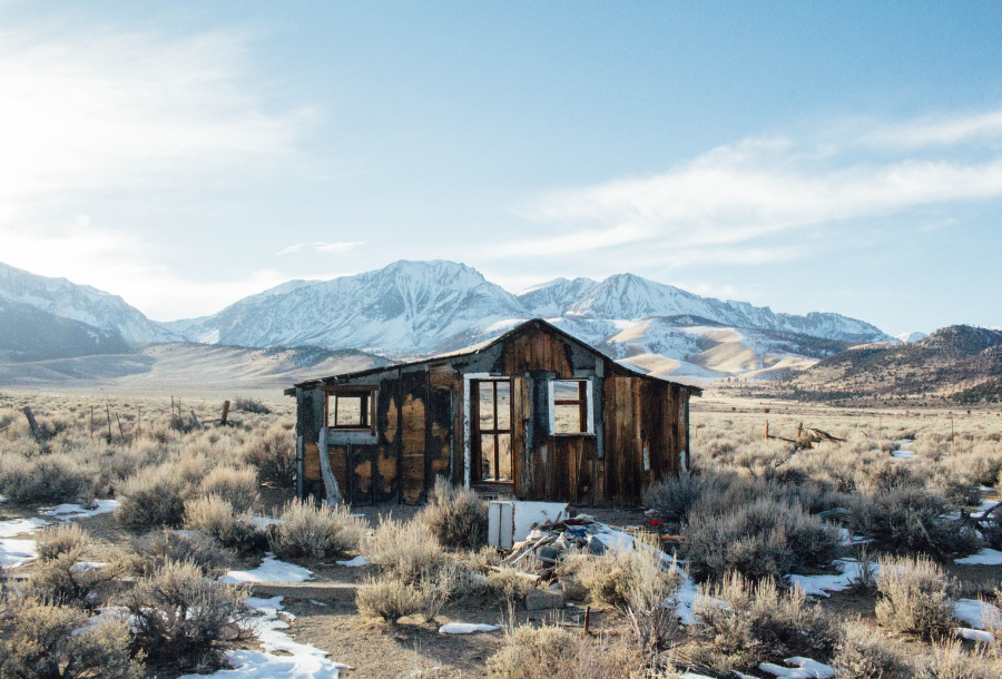 Picture of a rickerty old abandoned house in an open field, you are able to see in the far distance behind the house mountains that are covered in snow