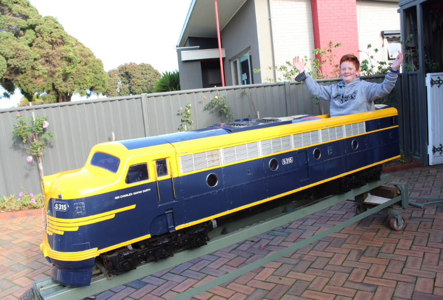 Picture of a young red head boy with his hands up in the air, enjoying riding in a stationary miniature railway carriage in a front bricked yard.