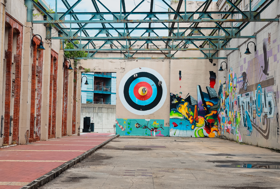 Picture of an old open warehouse with no roof just blue steel beams between the concrete walls - bright coloured graffiti artwork on the right hand size leading up to a huge painted white, black, blue, red and yellow bullseye