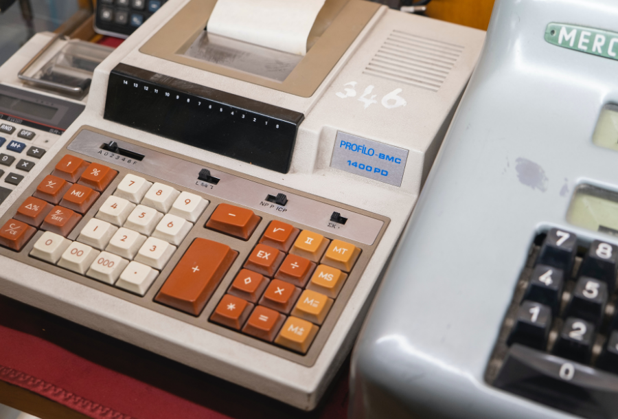 Picture of three calculators that are from an old era, a small one on the left, a big adding machine with orange, white and yellow buttons in the middle and a silver adding machine with black numbers on the right