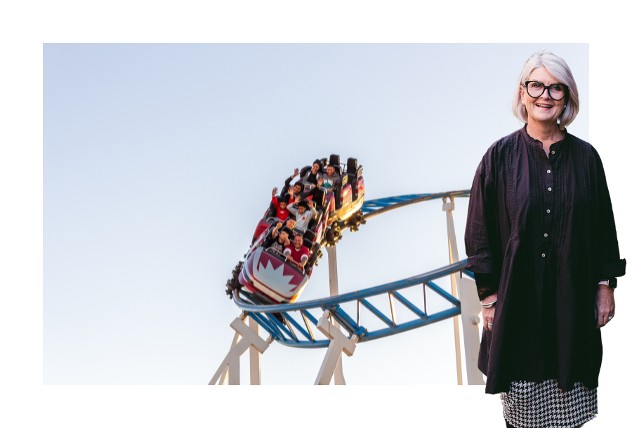 Pictured, Amanda Wilkens with a big smile on her face wearing a long black shirt with a black and white checked pencil skirt. In the background is a picture of a rollercoaster that is full of people, excited with their hands up in the air enjoying the ride. It's a clear sky day and the sun is shining brightly.