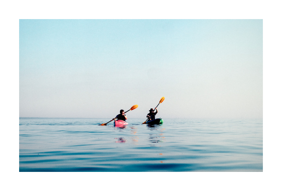 Pictured, two people canoeing across calm water, one in a bright orange canoe both using orange coloured paddles.