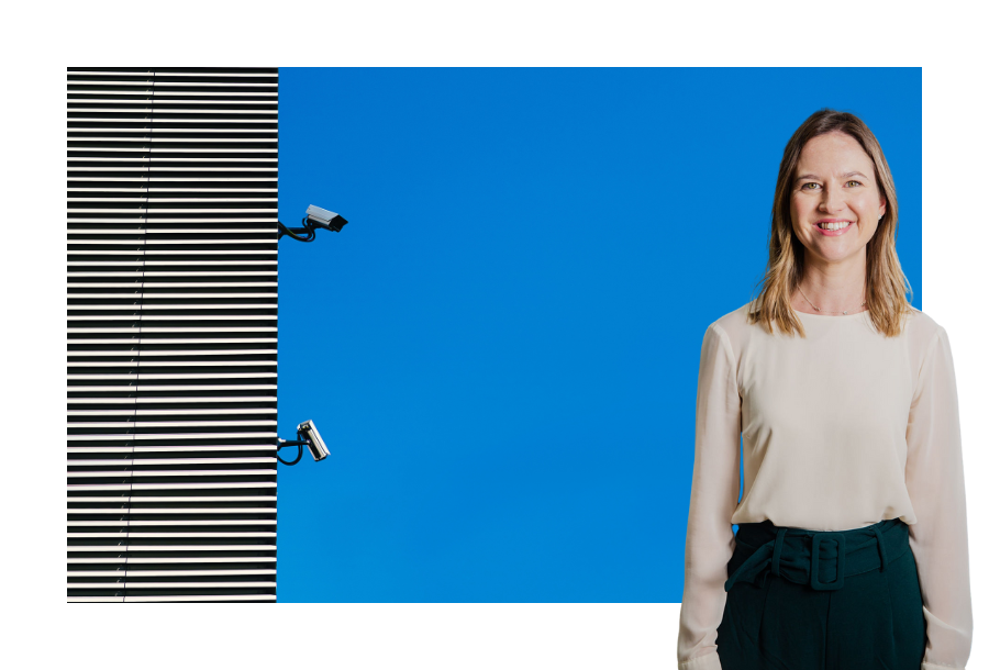 Pictured, Senior Lawyer Gabrielle Andersen standing in front of a picture of a building with security cameras against a clear blue sky.