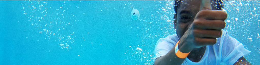 Picture of a young child with dark skin smiling under water wearing a white t-shirt and holding up his right hand giving the "thumbs up"