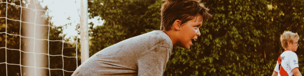 Picture of a young child playing soccer, wearing a dark grey t-shirt leaning over holding his knees, catching his breath in front of soccer nets