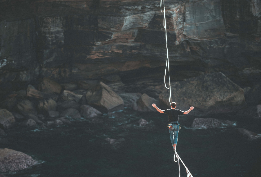 Pictured, a man wearing a dark coloured t-shirt with blue pants walking across a rope over a canyon surrounded by dark coloured rocks