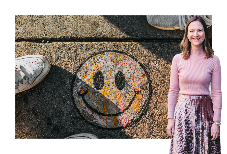 Pictured, Gabrielle Andersen wearing a long sleeve pink jumper with a pink velvet skirt. Standing in front of a stock picture of a painted outlined black smiley face on the pavement and two sets of white shoes on either side.