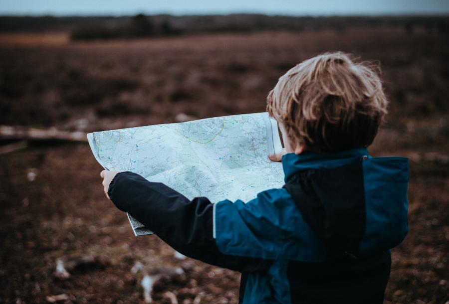 Young boy standing in an open field wearing a blue/black contract long sleeve jacket with a hood holding a map out in front of him in both hands