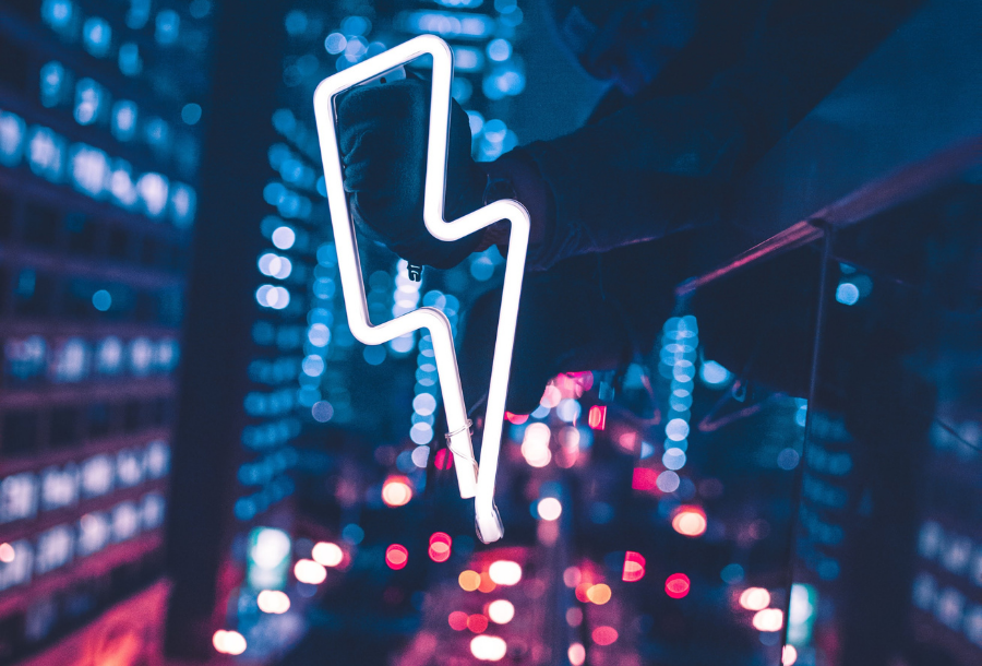 White neon lightning bolt in the foreground with a busy background of city lights and car break lights in the bottom of the background of the picture