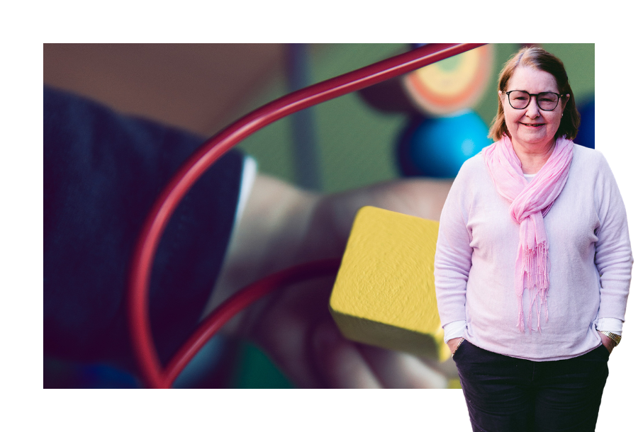 Pictured a close up photo of a child hand playing with a loop game with blocks. In the forefront Christine Friel stands wearing black pants, a long sleeve light pink coloured top and a pink scarf.