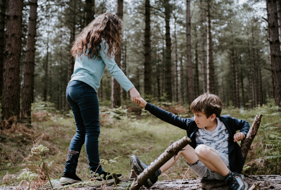 Young girl with curly brown hair standing on a fallen down tree branch holding out her hand to a younger boy in a white t-shirt with blue alligators to help him stand up