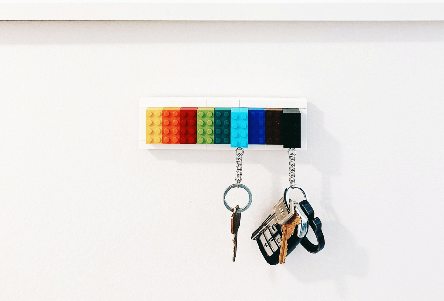 Two sets of keys hanging on a key rings placed against a white washed wall, the keys are hanging from a key holder made of yellow, orange, red, shades of green and blue lego pieces