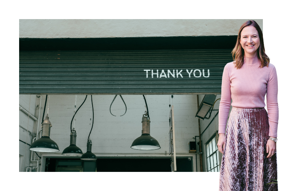 Pictured, Gabrielle Andersen wearing a dark pink velvet skirt and a long sleeve pink top. Standing in front of a stock picture of a half-opened dark green roller door, where you can see the word "THANK YOU" painted on and hanging light fittings from the roof.