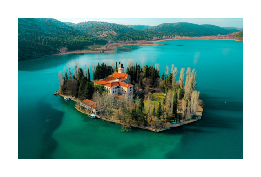 Pictured, a remote house surrounded by beautiful clear blue water on an island. Long tall green trees surround the house with a red roof