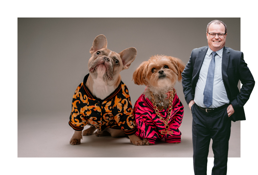 Pictured, Adam Wightman with a smile and his hands on his hips. In the background is a picture of two gorgeous dogs holding paws, dressed in bright sweaters and looking up.