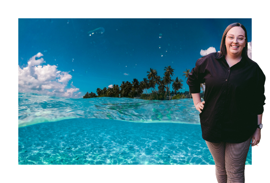 Pictured, Mekayla Lambert standing with one hand on her hip and a huge smile. Wearing a black long sleeve shirt and brown tweed colour pants, she is standing in front of a picture of a beautiful blue cleared water beach and sandy shores. There are palm trees in the background and a crystal clear blue sky.